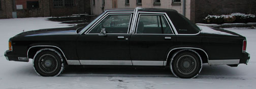 Black full-size four-door Ford sedan in profile on snow, wire wheel covers, brick building