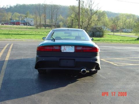 1993 Ford Probe - The rear of a teal car is shown parked on asphalt with a grassy field and buildings in the background. The license plate reads 'INDIANA 143…
