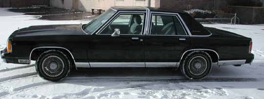 Black full-size Ford sedan in profile with wire wheel covers and chrome trim on snow