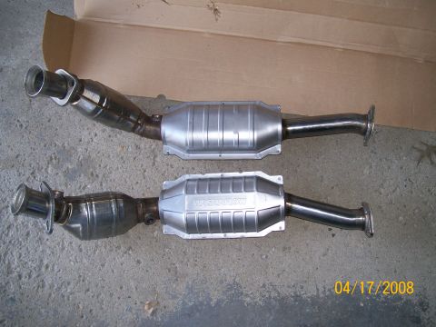 1995 Mercury Grand Marquis - Two catalytic converter exhaust assemblies laid side by side on a cardboard sheet over a concrete floor; the lower unit is a stainless Magn…