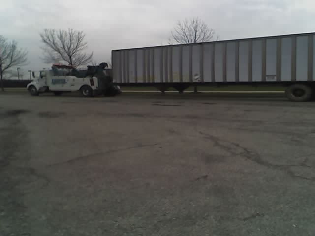 Tow truck is hooking up to a large trailer. Trees and overcast sky in background