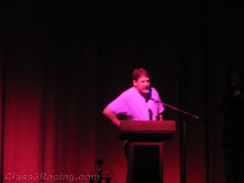 1990 Ford Bronco - man stands speaking at a wooden podium with a microphone under dim red stage lighting.