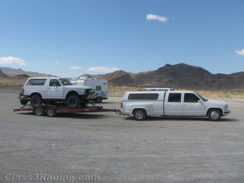 1990 Ford Bronco - white crew-cab dually pickup tows a flatbed trailer carrying a white-topped Ford Bronco across a desert lot.