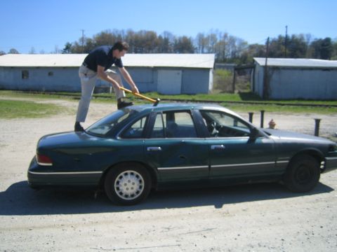 1985 Ford Crown Victoria LTD - Man prepares to strike the roof of a green sedan with a sledgehammer.