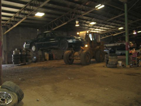 1985 Ford Crown Victoria LTD - A dark 1985 Ford Crown Victoria LTD is lifted by a forklift inside a warehouse, with other vehicles visible in the background.
