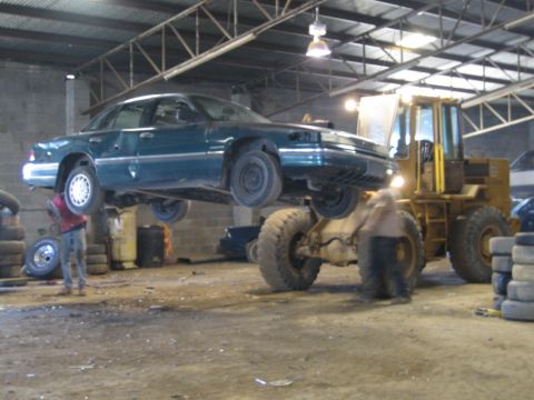 1985 Ford Crown Victoria LTD - A green sedan is being lifted by a yellow loader in what appears to be a junkyard.