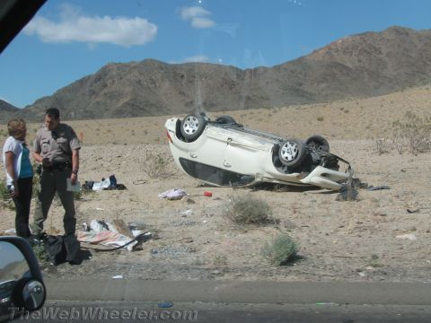 1990 Ford Bronco - white car rests upside down on its roof in the desert as a sheriff's deputy and a woman stand beside scattered debris.