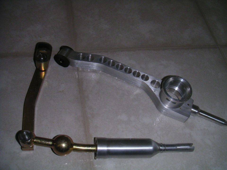 Shift knob and linkage parts on a tiled surface. Includes a brass ball joint and an aluminum lever with circular cutouts