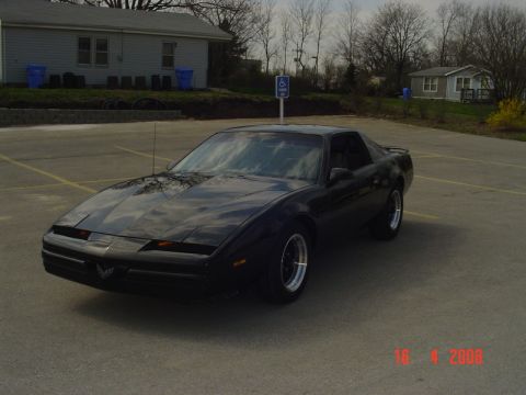 1989 Pontiac Firebird - A black Pontiac Firebird is parked in an empty lot with a building and trees in the background.