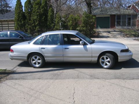 1996 Ford Crown Victoria - A silver 1996 Ford Crown Victoria is parked on the side of the road with a house and trees in the background.