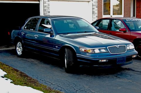1985 Mercury Grand Marquis - A dark blue sedan is parked in a driveway in front of a garage, with another car visible behind it.