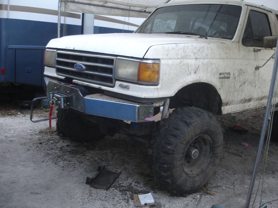 White Ford Bronco with winch bumper and large tires, covered in mud. Front 3/4 view