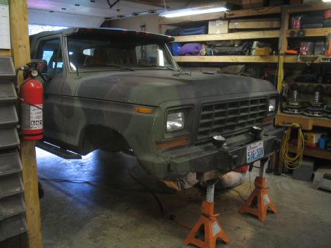 1979 Ford Bronco - Camouflage Ford Bronco undergoing work, supported by jack stands in a garage. The vehicle appears to be from the late 1970s.