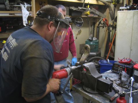 1979 Ford Bronco - Two men work on a metal component in a cluttered workshop setting, with tools and equipment visible in the background.