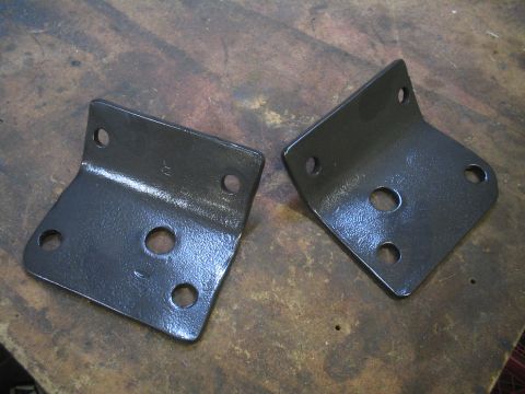1979 Ford Bronco - Two metal brackets with various bolt hole configurations are shown on a workbench.
