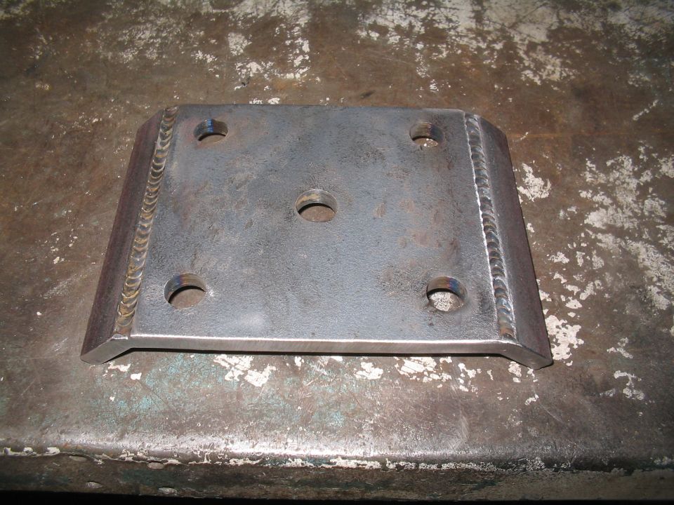 Metal plate with four outer bolt holes and one central hole, showing visible weld marks along the edges