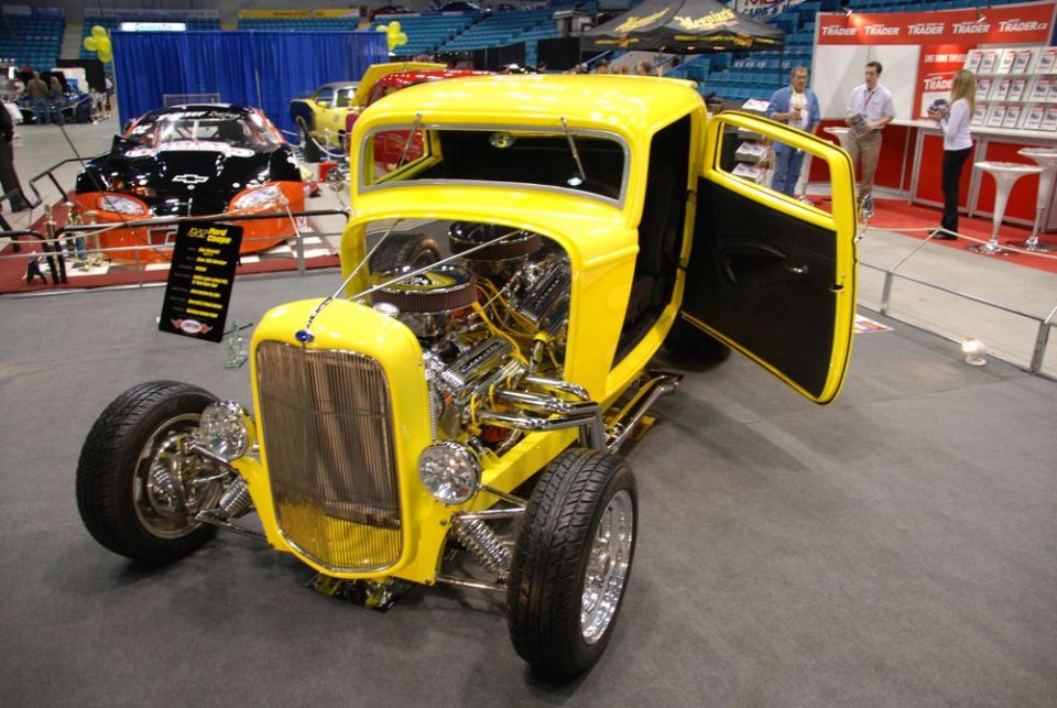 Yellow hot rod with open driver's door and exposed V8 engine, parked indoors. Another car is visible in the background
