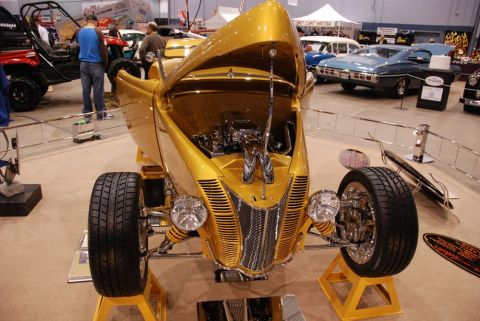 1996 Ford Bronco - A custom gold hot rod is displayed with its hood open, revealing the engine, at an indoor car show.