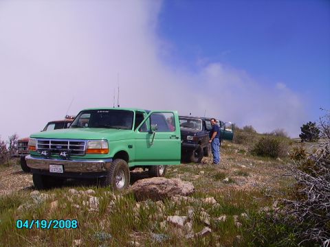 1990 Ford Bronco - Several trucks are parked on a grassy, rocky hillside under a partly cloudy sky.