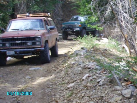 1990 Ford Bronco - Two off-road vehicles navigate a rocky dirt path through a wooded area.