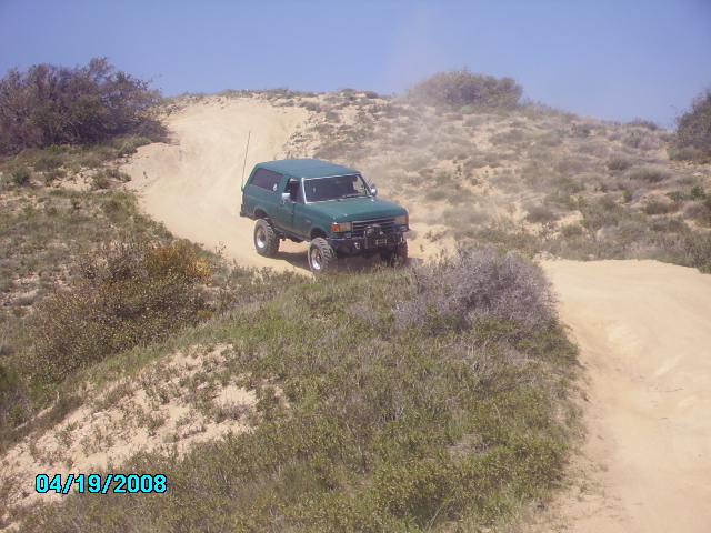 Green 1990 Ford Bronco driving up a sandy hill with dust kicking up