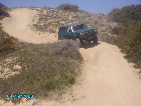 1990 Ford Bronco - A green SUV navigates a sandy track through a coastal landscape.