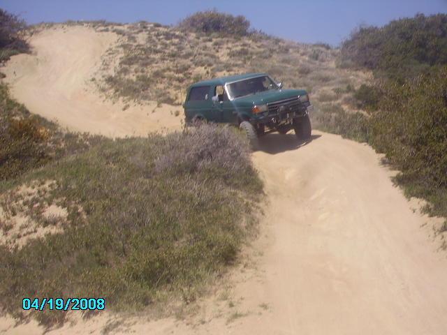 Green SUV driving on a sandy dirt road with brush and dunes