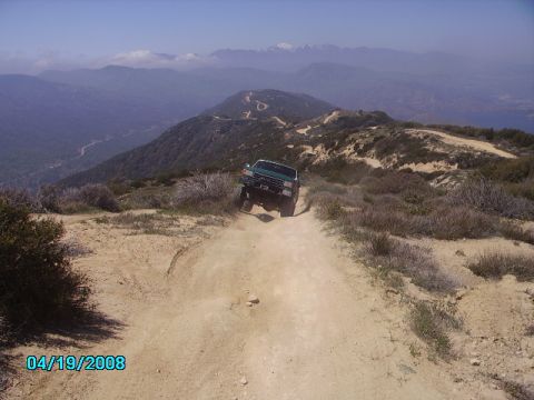 1990 Ford Bronco - A green SUV navigates a dusty mountain trail with scenic vistas and snow-capped mountains in the distance.