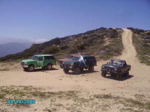 1990 Ford Bronco - Four off-road vehicles are parked on a dirt road on a sunny day. The green SUV on the left resembles a 1990 Ford Bronco.