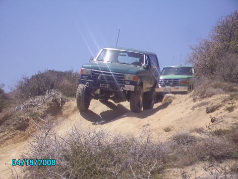 1990 Ford Bronco - Two green Ford Broncos navigate a sandy, off-road trail under a clear blue sky.