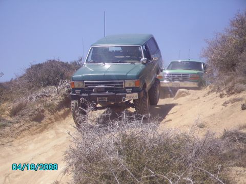 1990 Ford Bronco - Two green Ford Broncos navigate a dusty off-road trail under a clear blue sky.