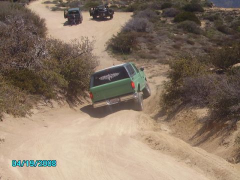 1990 Ford Bronco - A green 1990 Ford Bronco navigates a steep dirt path off-road, with two other vehicles visible in the background.