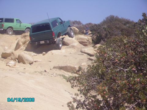 1990 Ford Bronco - Two green Ford Broncos navigate a rocky off-road trail under a clear blue sky.