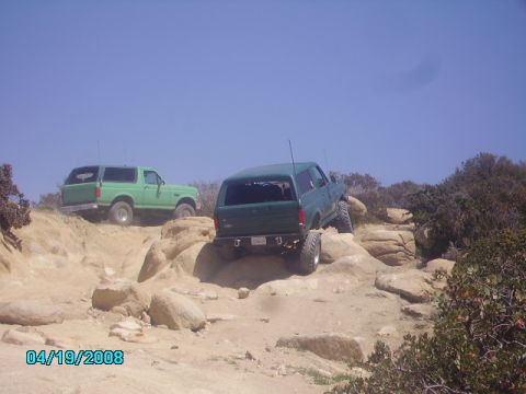 1990 Ford Bronco - Two green Ford Broncos tackle a challenging rocky trail under a clear blue sky.
