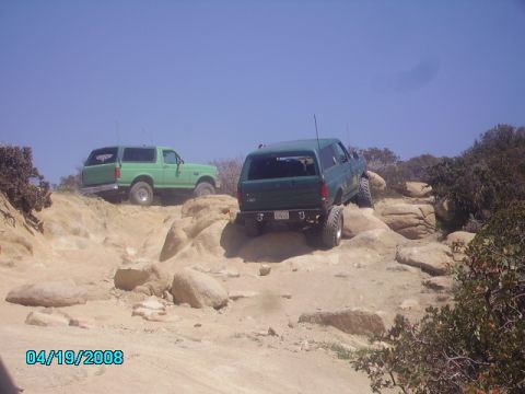 1990 Ford Bronco - Two green Ford Broncos tackle a challenging rocky terrain on a sunny day.