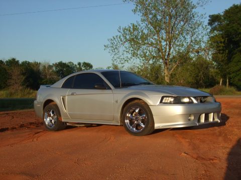 1999 Ford Mustang - A silver Ford Mustang is parked on a dirt surface under a clear sky, showing its front 3/4 profile.