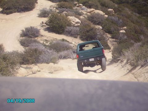 1990 Ford Bronco - A green Ford Bronco navigates a dusty off-road path with scrub brush and rocks.