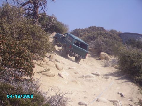 1990 Ford Bronco - 1990 Ford Bronco navigates a steep, rocky off-road path under a clear blue sky.