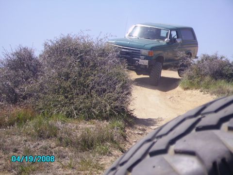 1990 Ford Bronco - A green Ford Bronco navigates a dusty, off-road trail under a clear blue sky.