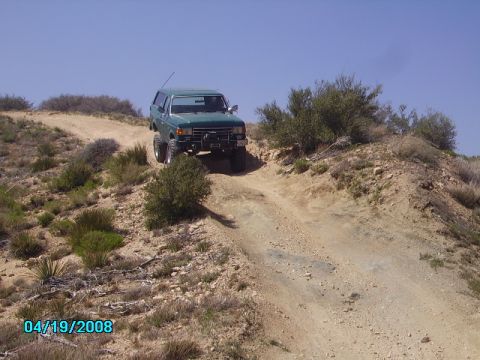 1990 Ford Bronco - A green Ford Bronco navigates a dusty, off-road trail under a clear blue sky.