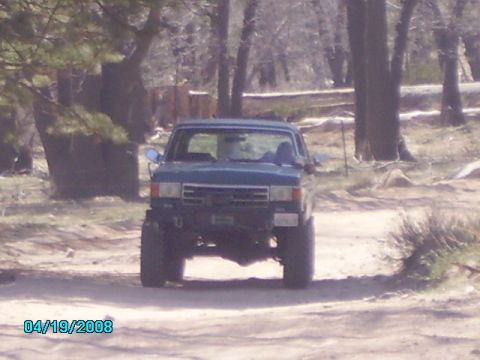 1990 Ford Bronco - Front view of a blue 1990 Ford Bronco driving on a dirt road surrounded by trees.