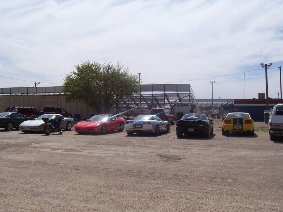 Several sports cars, including a red Corvette and a silver Corvette, parked in a lot with a motorcycle in front. A yellow Mustang is also visible