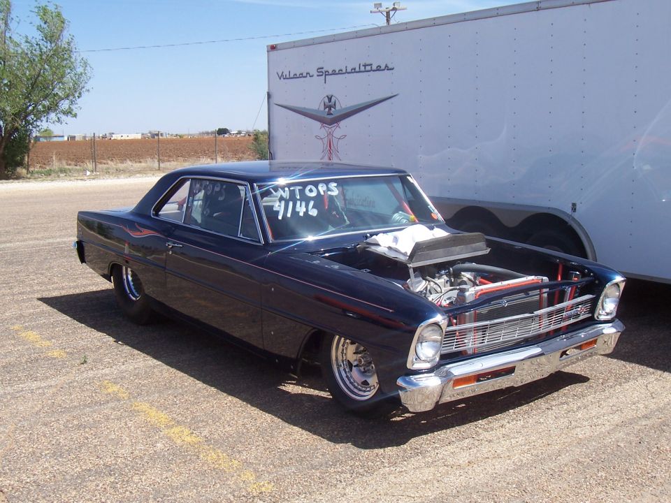 Dark blue coupe with racing modifications, hood open, parked next to a white trailer