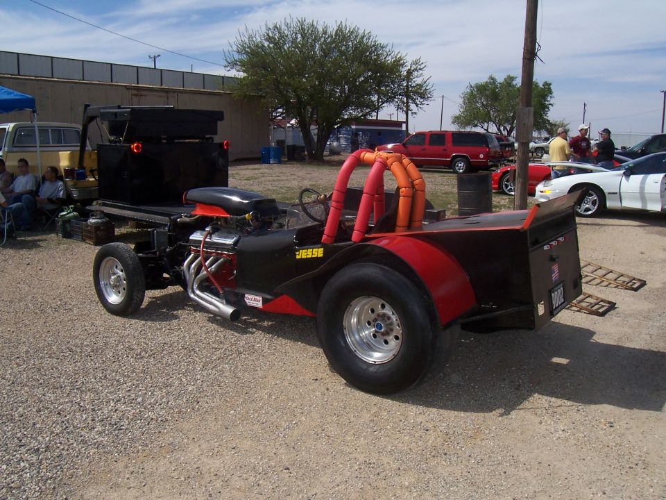 Dragster with a flatbed trailer, engine exposed, red and black paint, "JESSE" on the side. Other cars and people in background