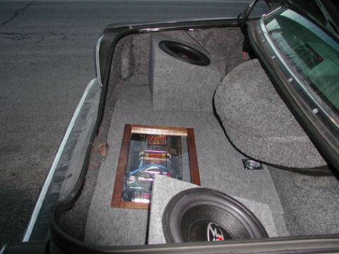 1988 Ford LTD Crown Victoria - Open trunk of a sedan with a custom gray-carpeted stereo installation: two subwoofers mounted in angled enclosures and a wood-trimmed clear…