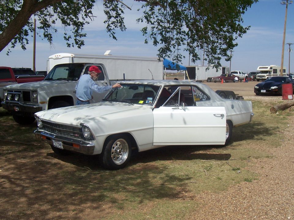 White 1966-1967 Chevrolet Nova coupe with driver's door open, man in red hat leaning in. Pickup truck behind it