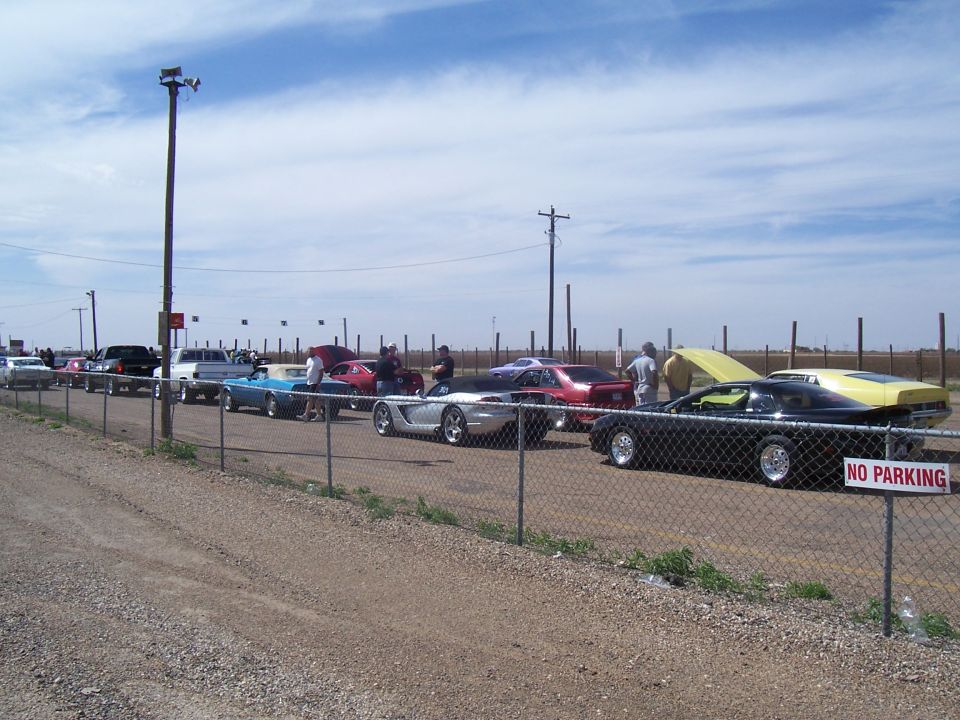 Several cars are parked in a line at what appears to be a drag strip, with a fence in the foreground