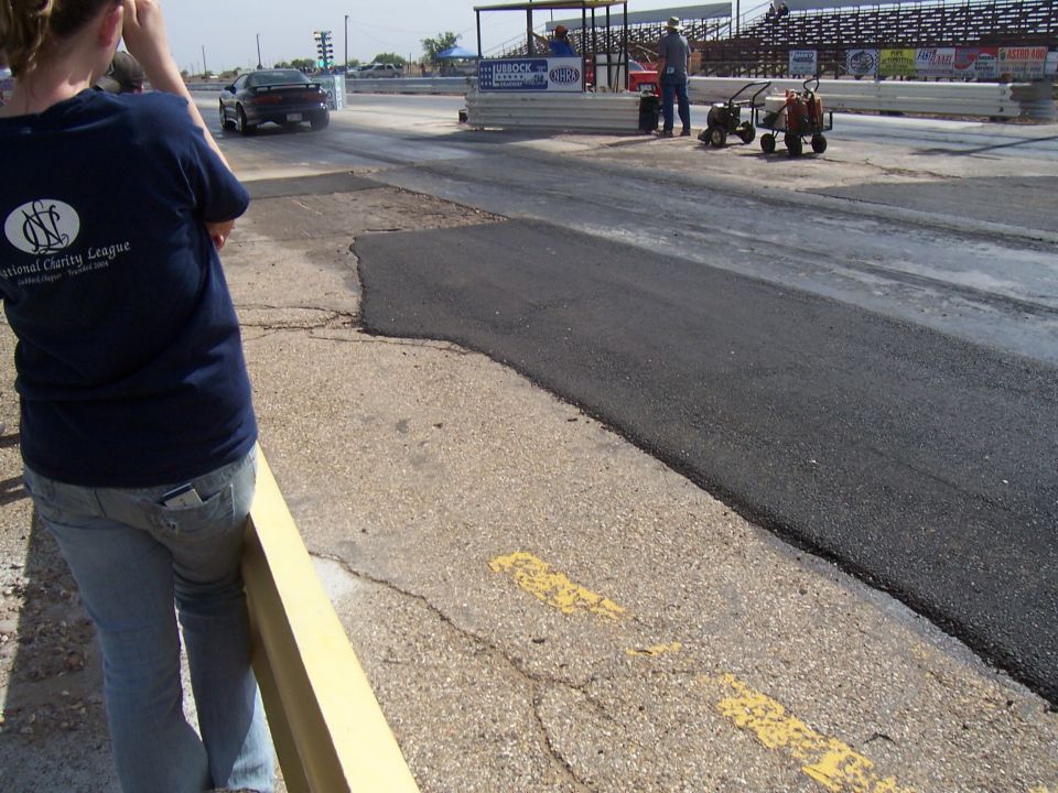 Drag strip with a car accelerating away, people and equipment near the start line