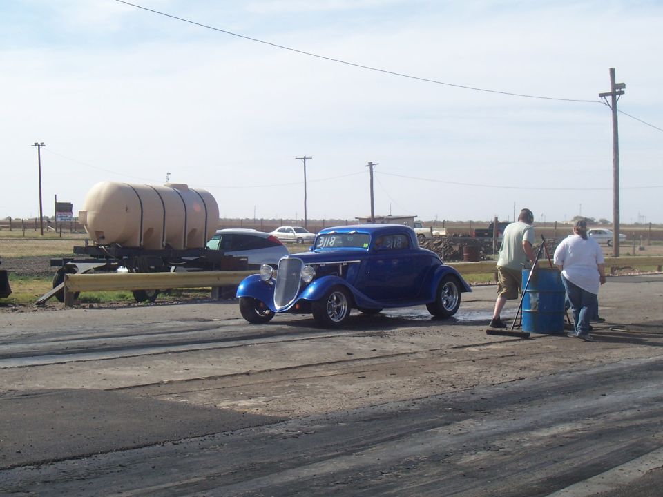Blue hot rod coupe with chrome grille and headlights on a drag strip, with two people and a barrel nearby