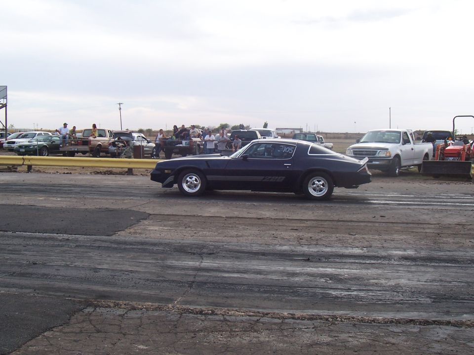 Dark blue coupe with racing stripes and number 105 on door, parked on a drag strip with other vehicles and spectators in background